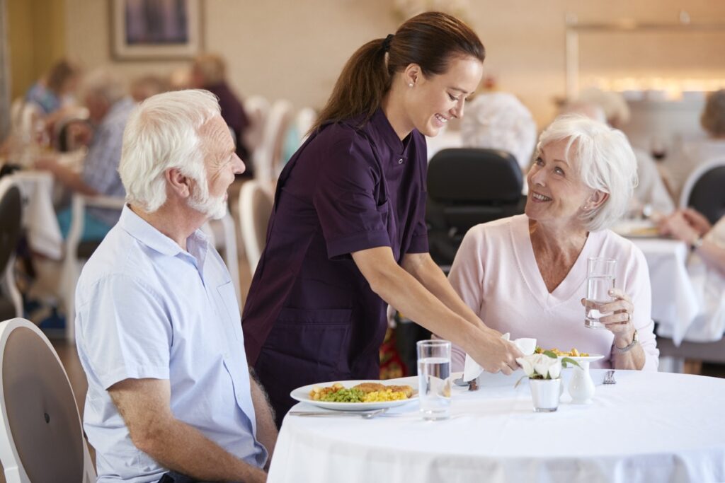 Senior Couple Being Served With Meal By Carer In Dining Room Of Retirement Home