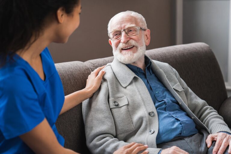 Young friendly african female caregiver talking chatting to happy senior man in hallway of nursing home. Picture of smiling nurse assisting senior elderly man